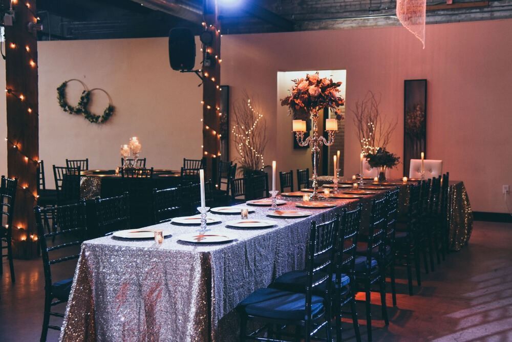 Long dining table set for a formal event, with shimmering tablecloth and black chairs, candles and flowers as centerpieces.