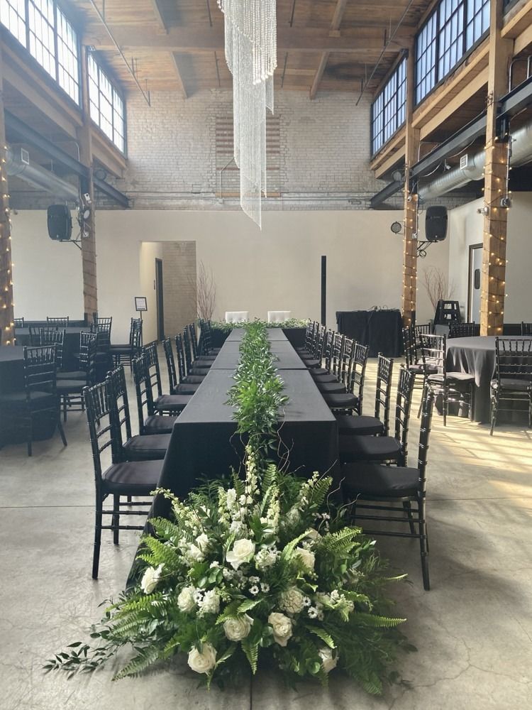 Long dining table with black tablecloth and chairs, floral centerpiece, in a bright, industrial-style venue.