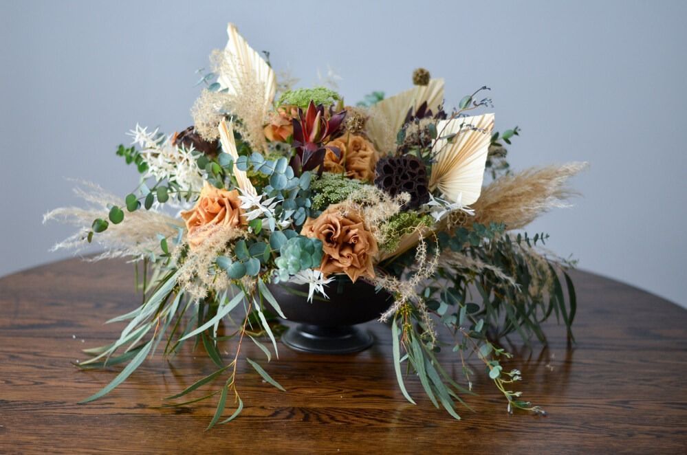 Floral centerpiece in a black bowl on a wooden table, featuring dried flowers in shades of beige, brown, and green.