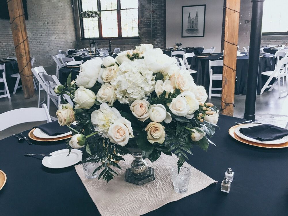 Elegant white floral centerpiece on a dark blue table setting, with a backdrop of a venue with chairs and tables.
