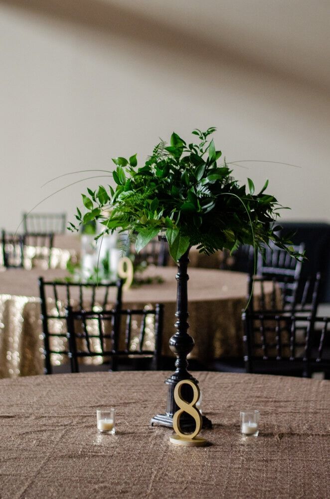 Wedding reception centerpiece: Green foliage, gold table number 8, brown tablecloth, and black chairs.