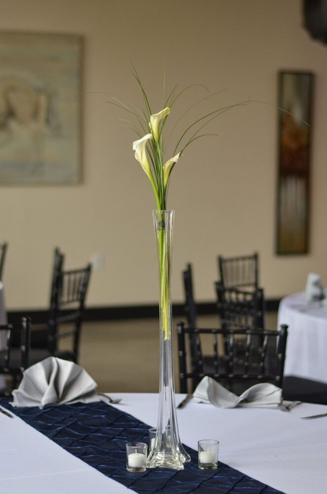 Tall glass vase with calla lilies on a white tablecloth; navy runner, candles, and napkins.