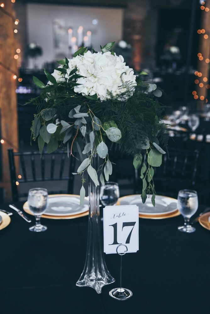 Elegant centerpiece with white flowers, greenery, and a table number on a black tablecloth.