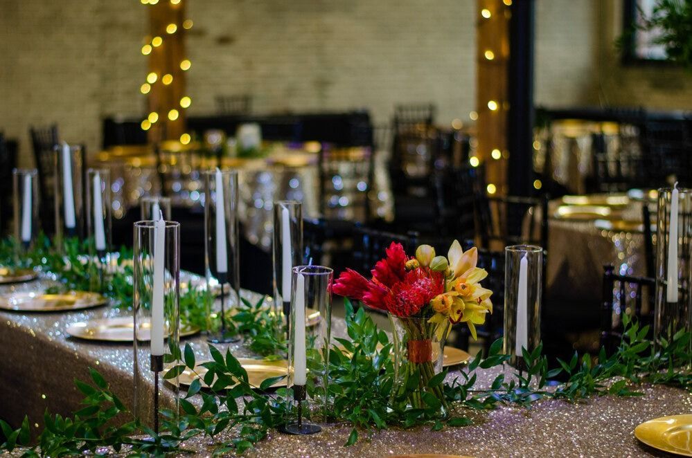 Long table set for a formal event, featuring flowers, candles, gold plates, and greenery.