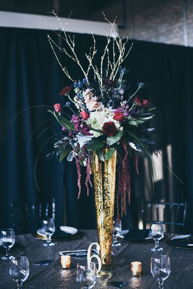 Tall gold vase with flowers on a table set for a formal event.  Dark blue backdrop with table setting and candles.