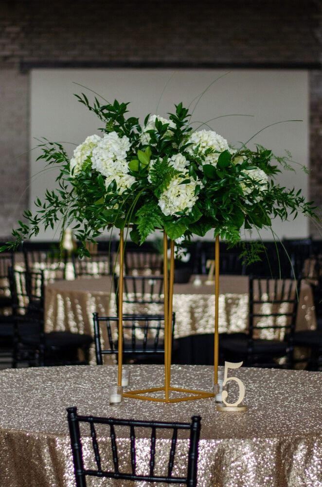 Floral centerpiece on gold stand atop a sequined table, black chair in foreground.