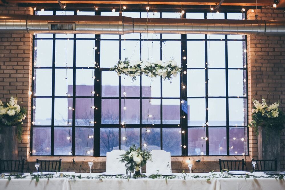 Wedding reception table set in front of a large window with city view. Decorated with flowers and lights.
