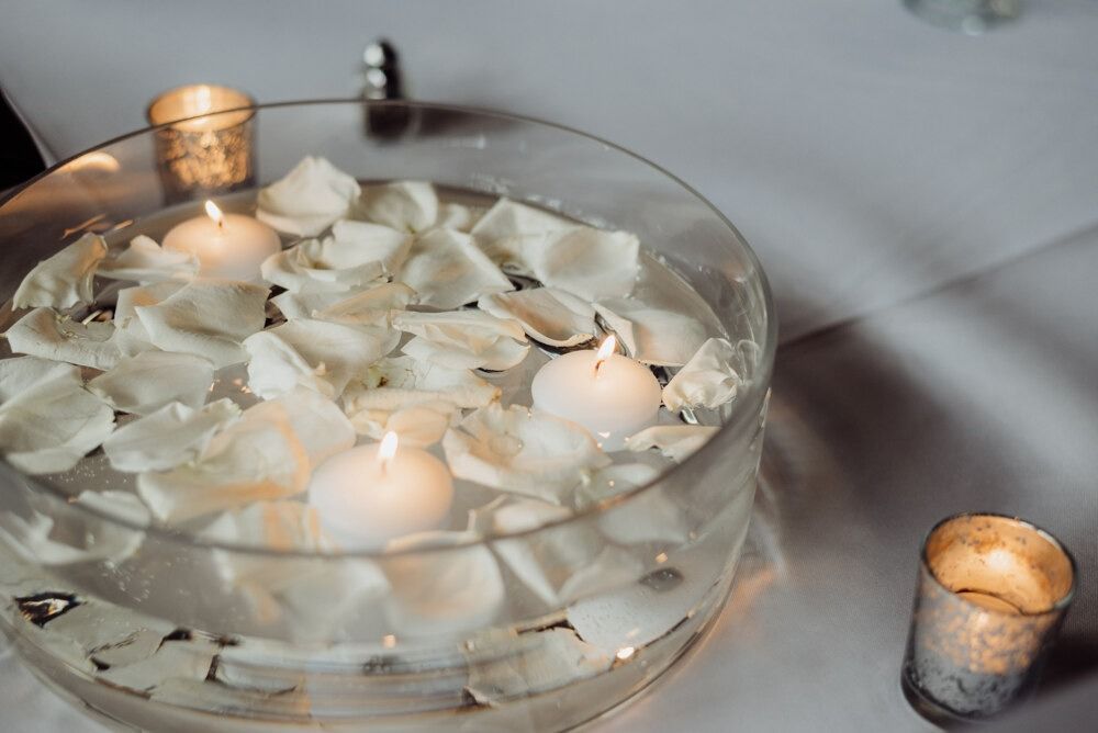 A clear glass bowl with floating candles and white flower petals on a table.