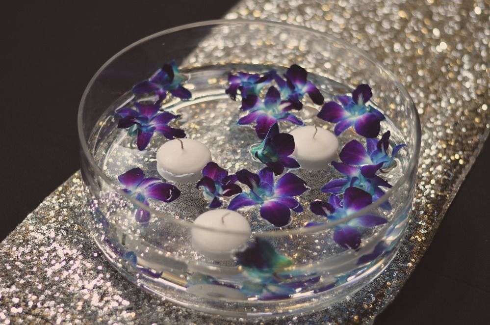 Floating candles and purple orchids in a glass bowl on a sparkly silver surface.