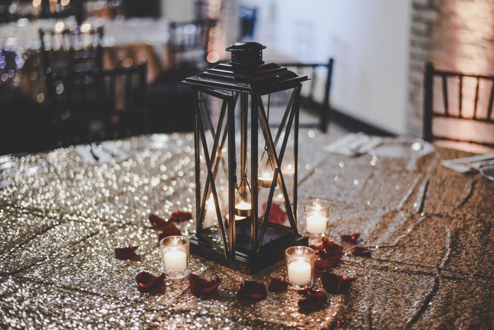 Black lantern centerpiece with candles and red rose petals on a sequined tablecloth.