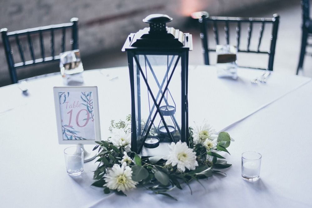 Wedding table centerpiece: black lantern, white flowers, greenery, table number.