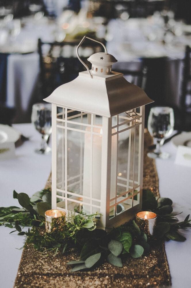 White lantern centerpiece on a gold sequin runner, with greenery and candles, set on a white tablecloth.