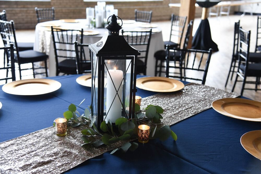 Table setting with black lantern, gold charger plates, and silver runner on blue tablecloth.