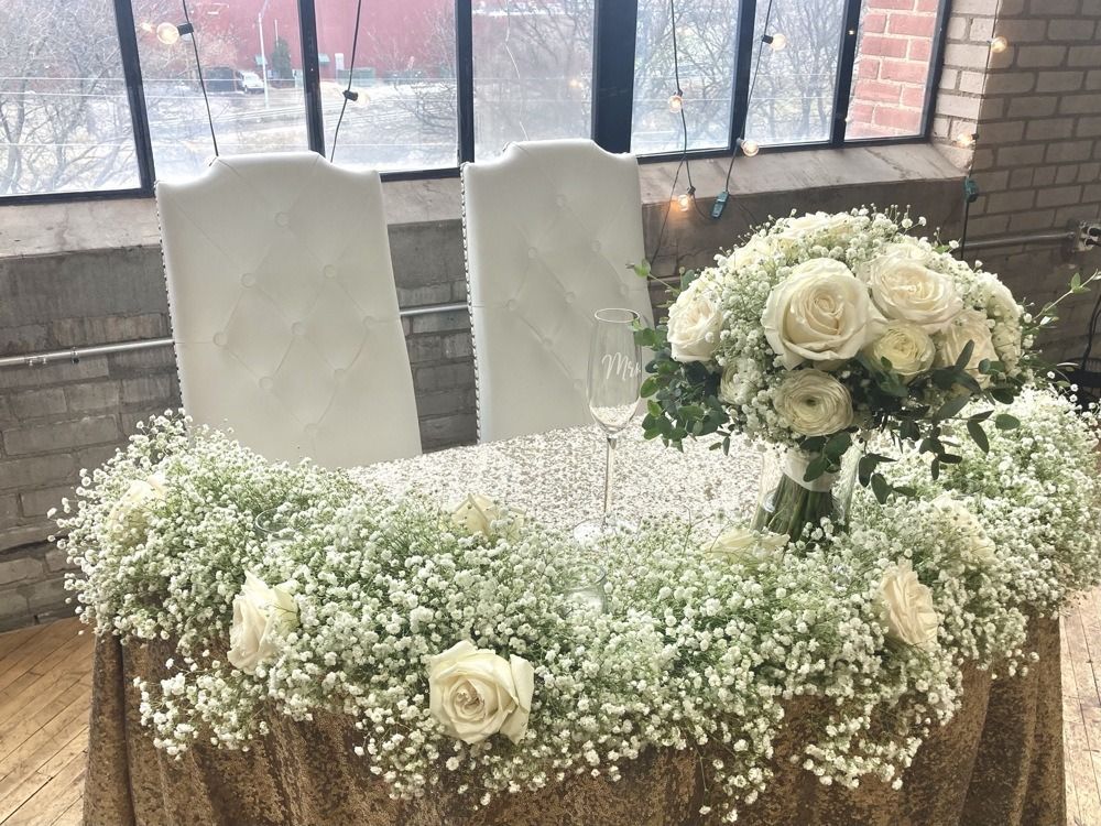 Wedding table decorated with baby's breath and white roses; two white chairs sit behind the table.