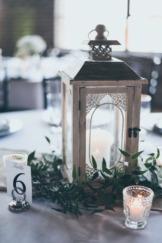 White lantern with lit candle, greenery, and table number 6 on a wedding table setting.