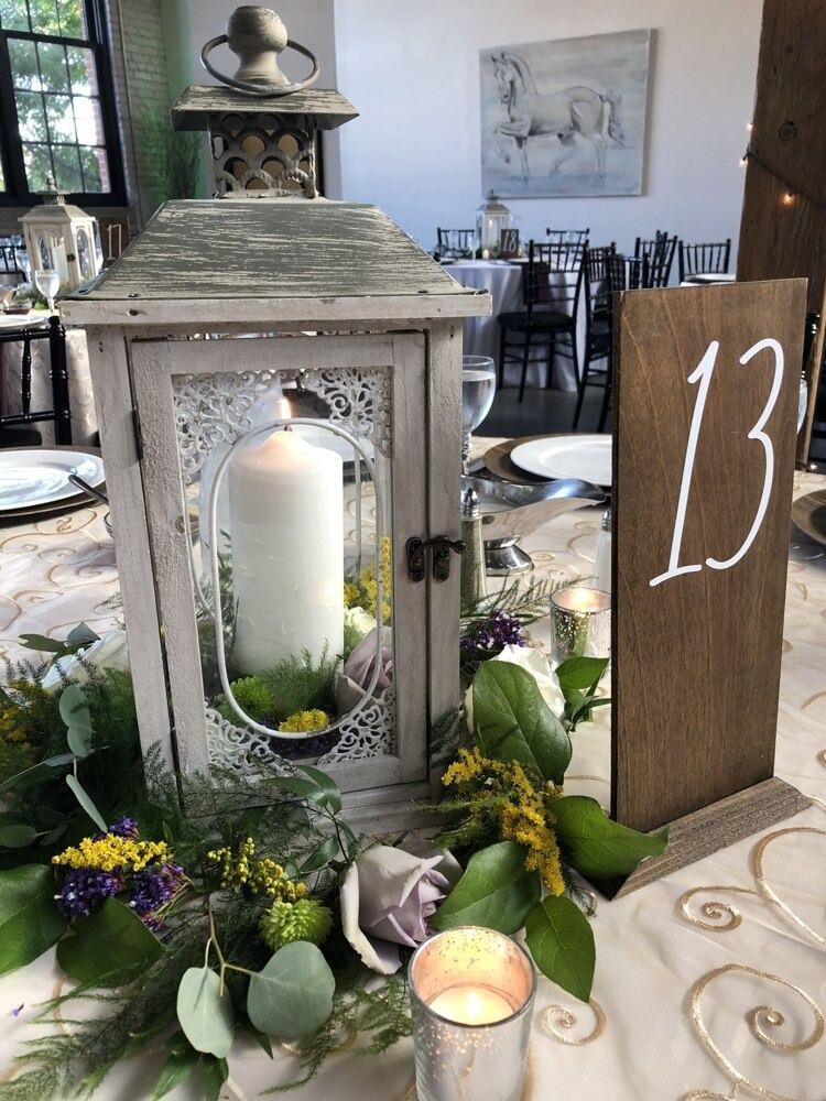 Centerpiece with lantern, candle, greenery, and table number 13 on a decorated table.