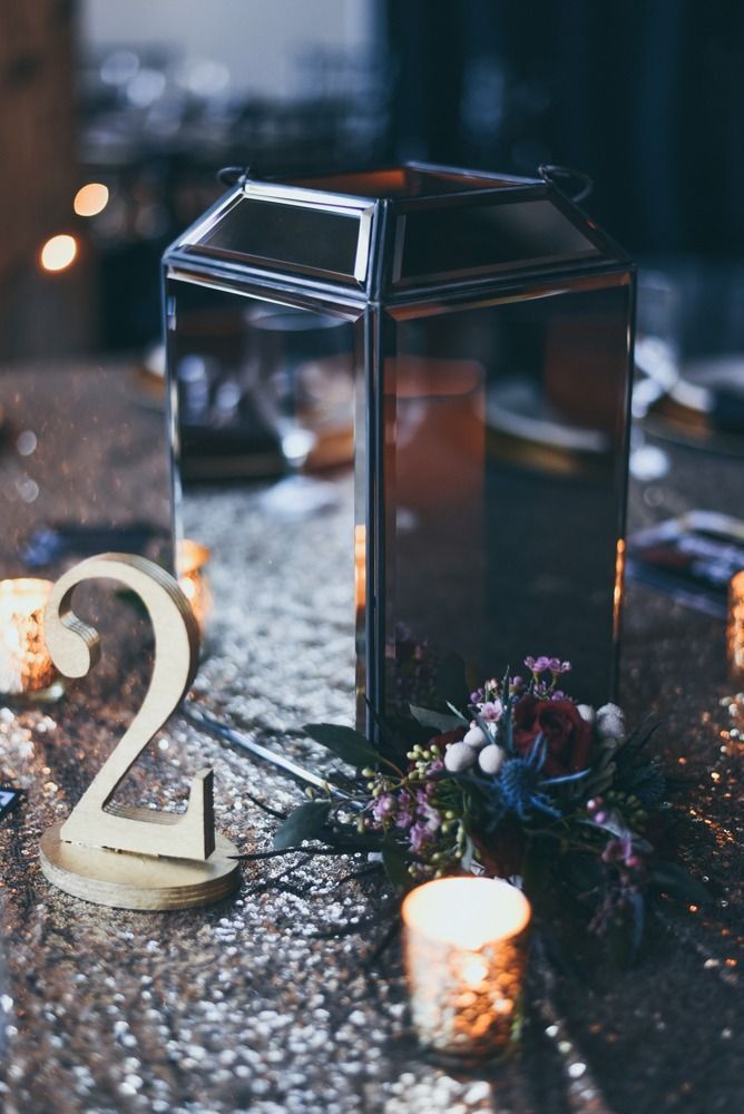 Metallic lantern centerpiece on a sequined tablecloth, with a table number and flowers. Lit candles in the foreground.