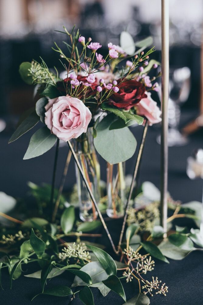 Floral centerpiece with pink and burgundy roses, greenery in gold geometric stand on dark table.