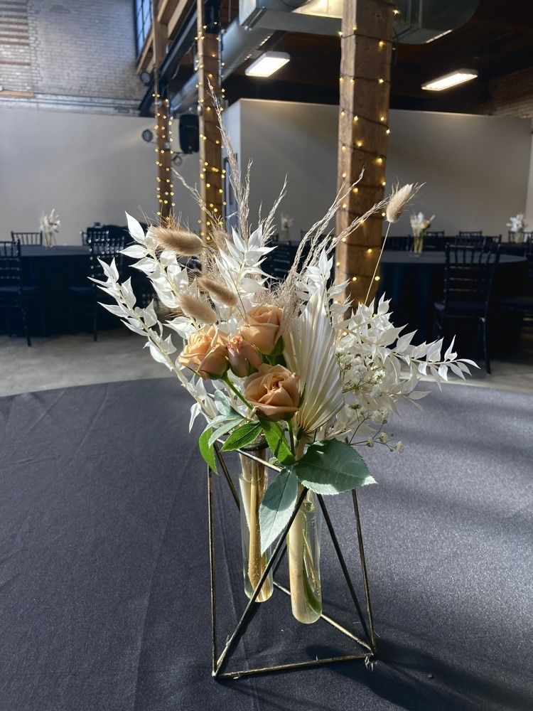 Floral centerpiece with dried beige and white flowers in a gold geometric stand, on a table.