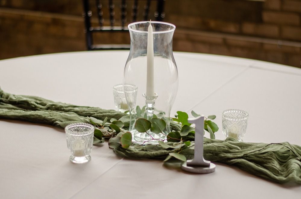 Table setting with a white tablecloth, centerpiece with greenery, candles, and a table number.