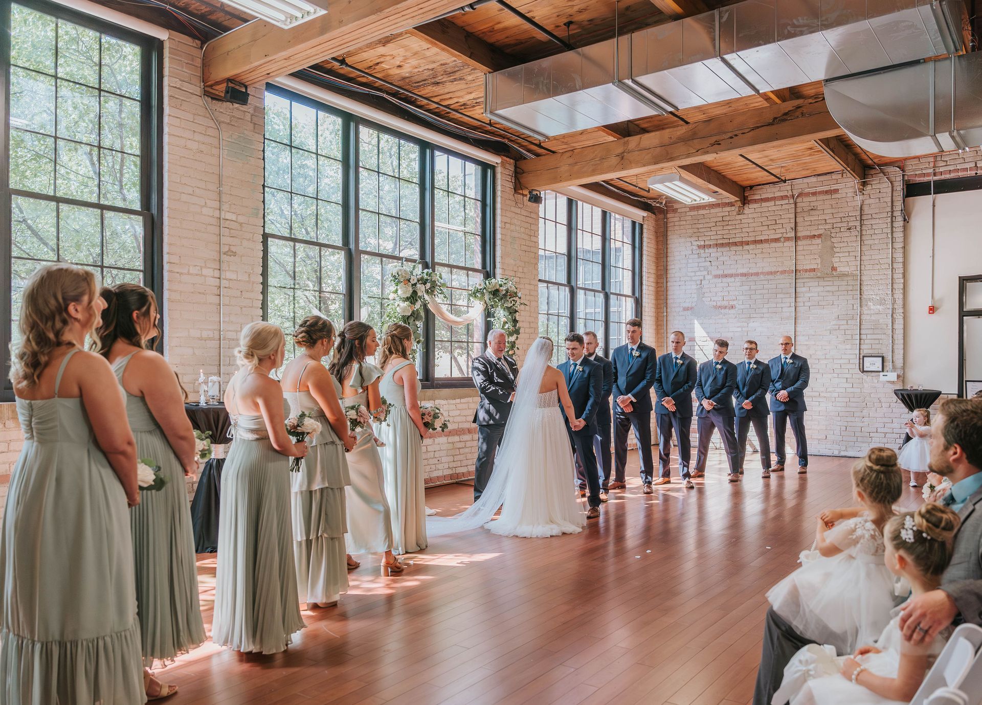 Wedding ceremony in a large room with windows; bride and groom at altar; wedding party stands on either side.
