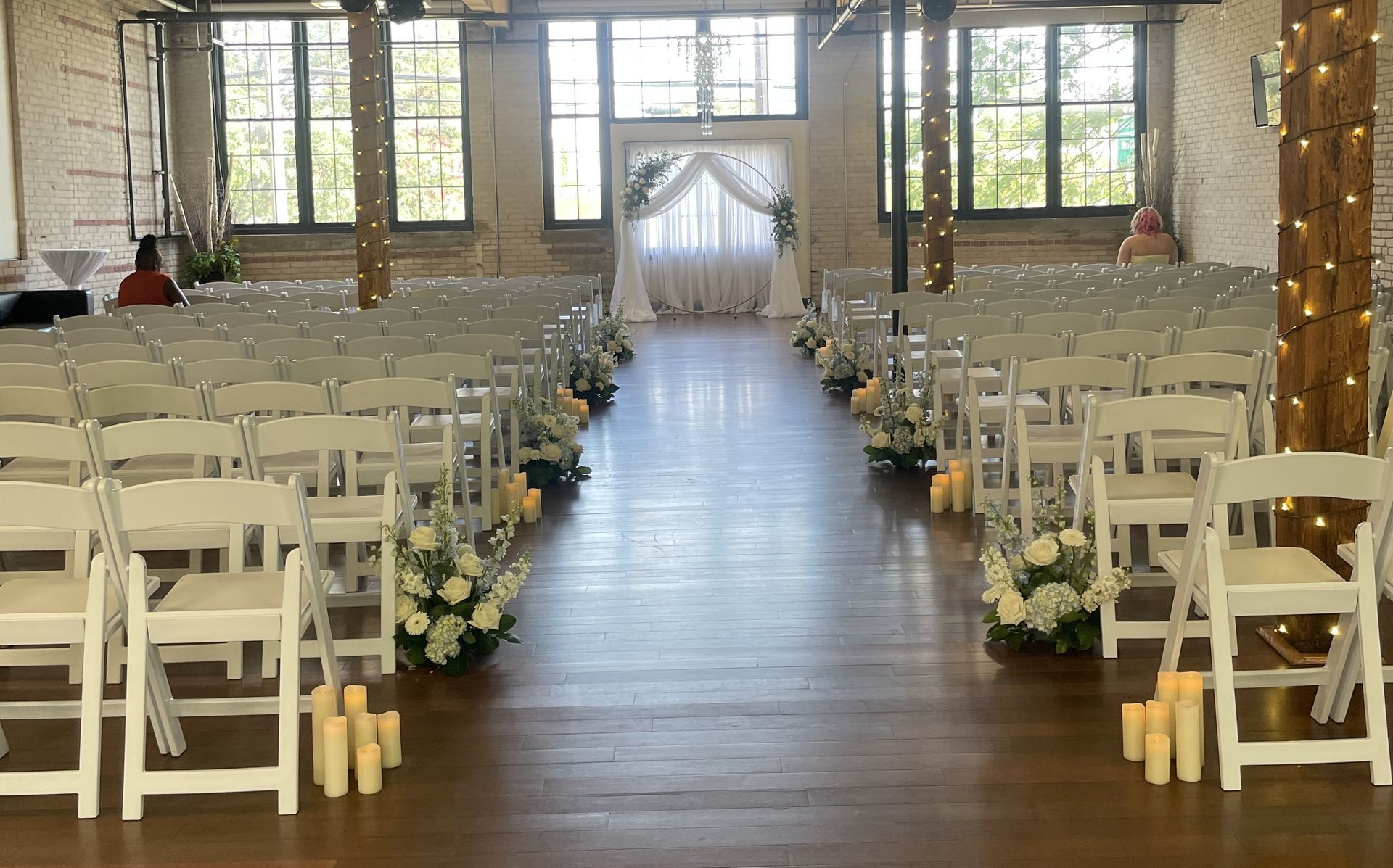 Wedding ceremony setup, white chairs lining the aisle, floral arrangements, string lights, and draped archway.
