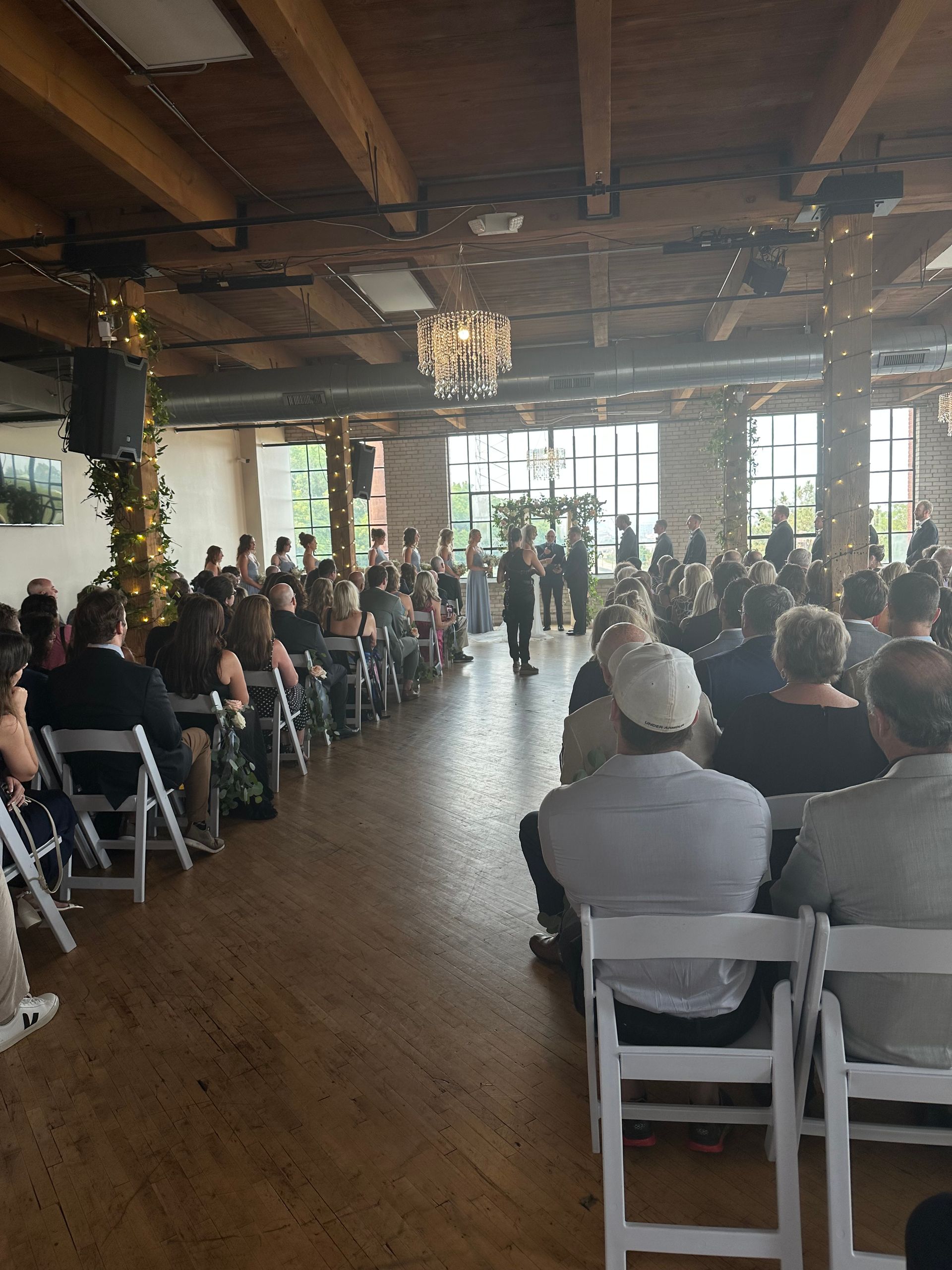 Wedding ceremony in a rustic venue, guests seated, couple at the altar. Wooden beams and large windows.