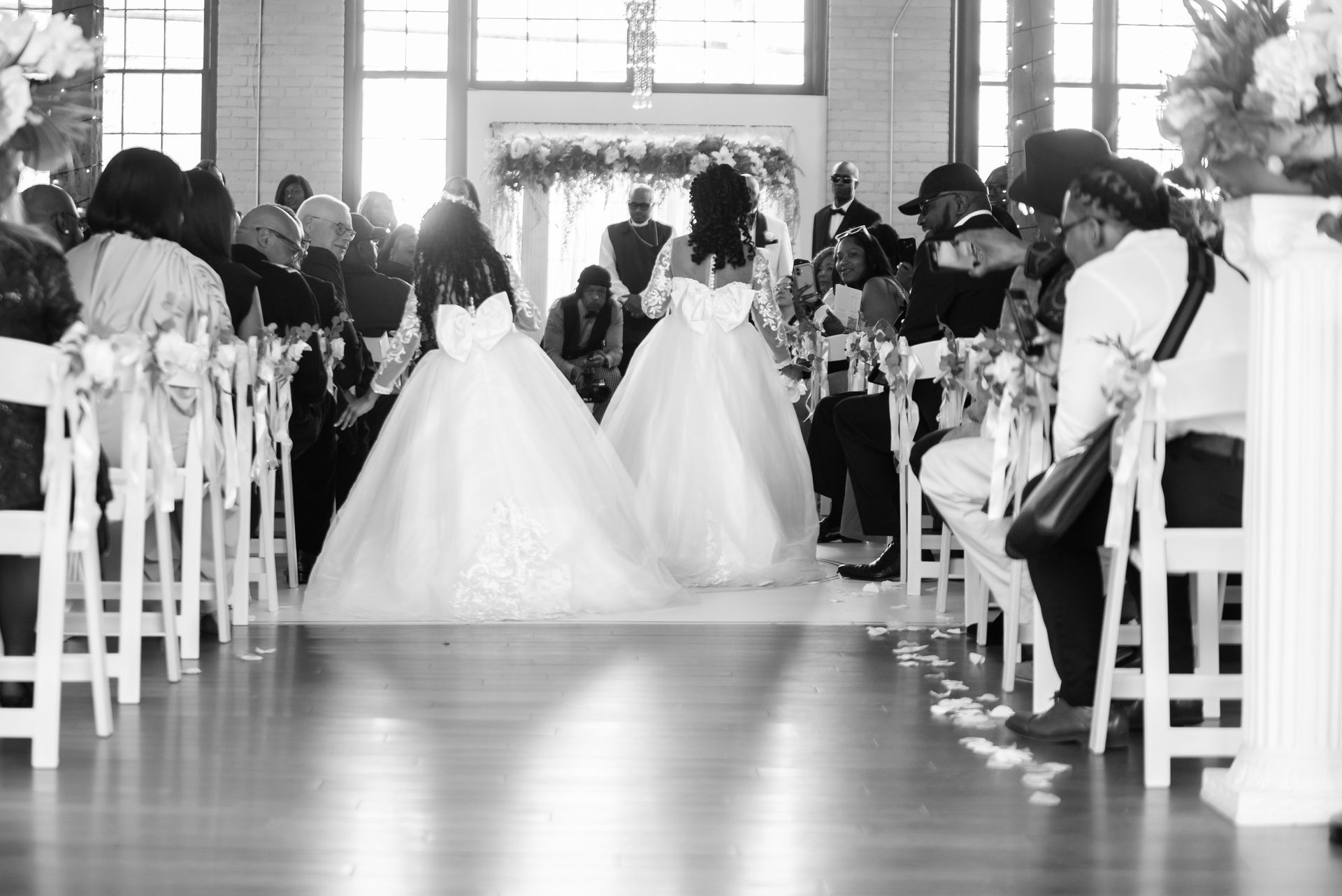 Two brides in white gowns walk down the aisle at a wedding ceremony. Guests watch from pews.
