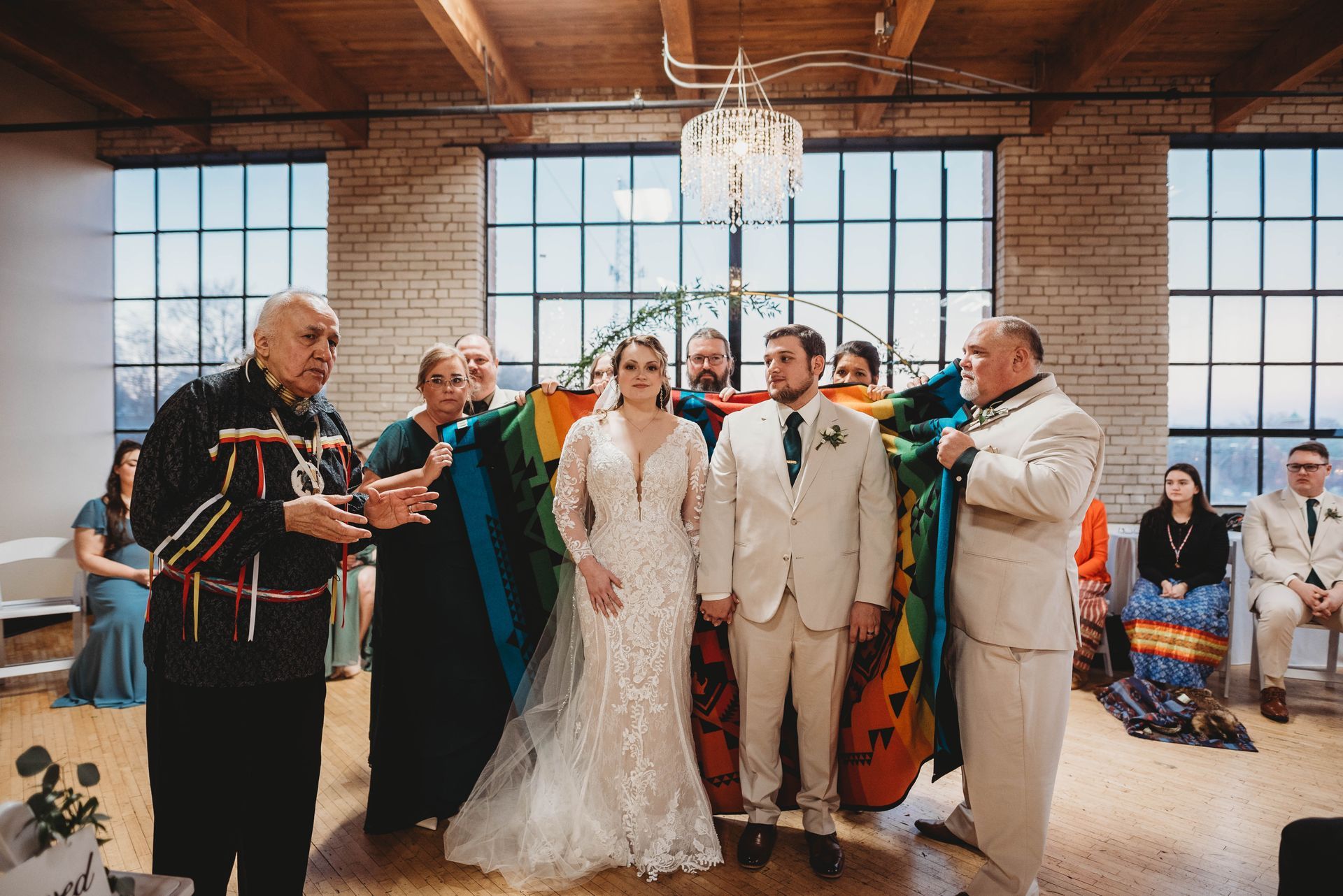 Wedding ceremony: Couple stands under a rainbow blanket. Officiants speak with guests looking on.