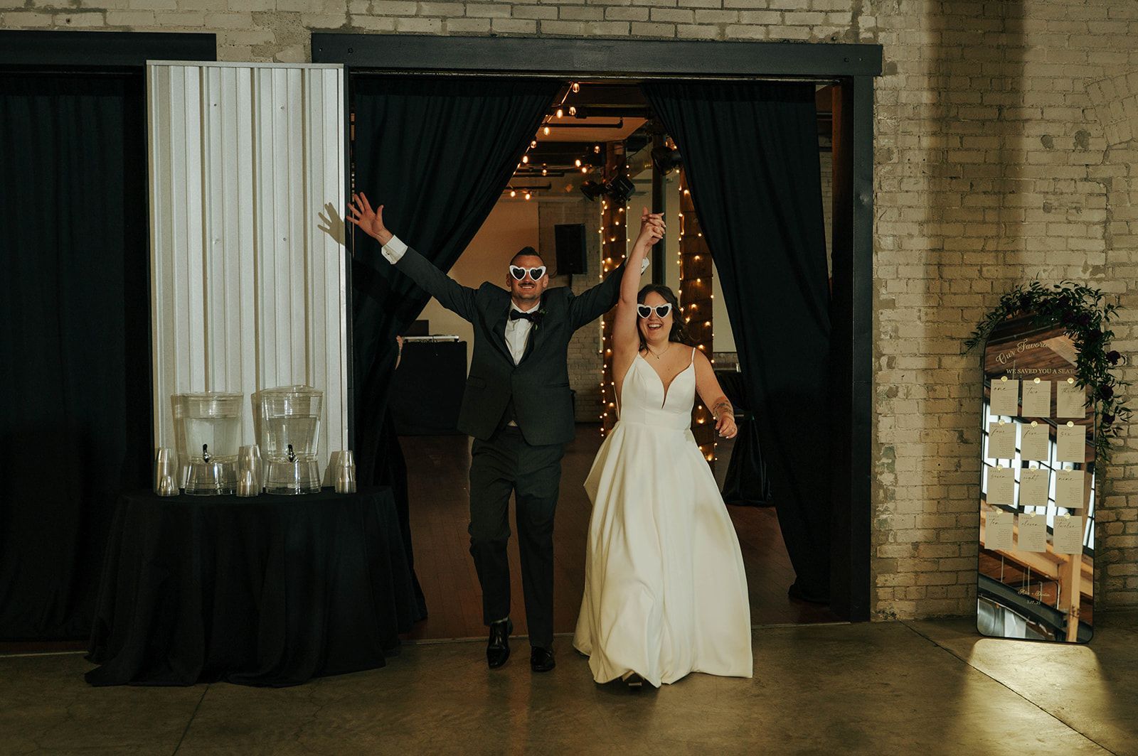 Bride and groom in sunglasses jump through doorway into reception. Black curtains, white dress, green foliage, bright interior.