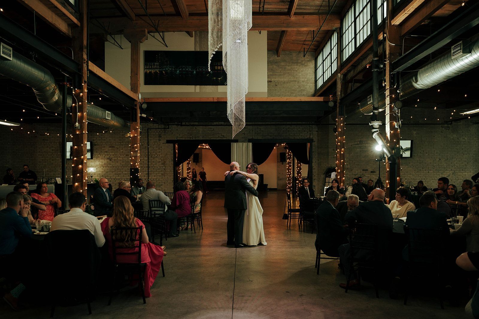 Bride and groom dance at reception in a large hall, surrounded by guests at tables. String lights and decor.