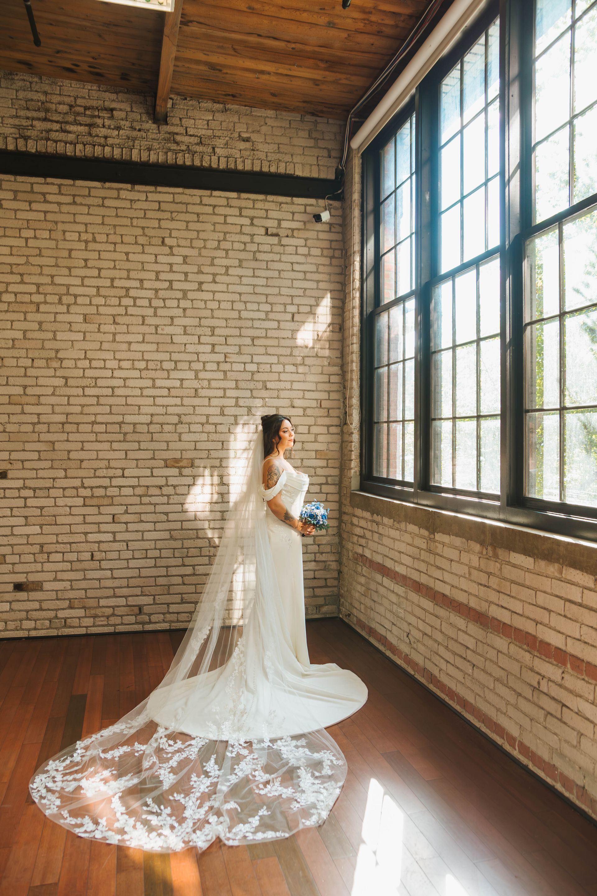 Bride in white dress, veil, and bouquet, standing by a large window in a room with exposed brick walls.