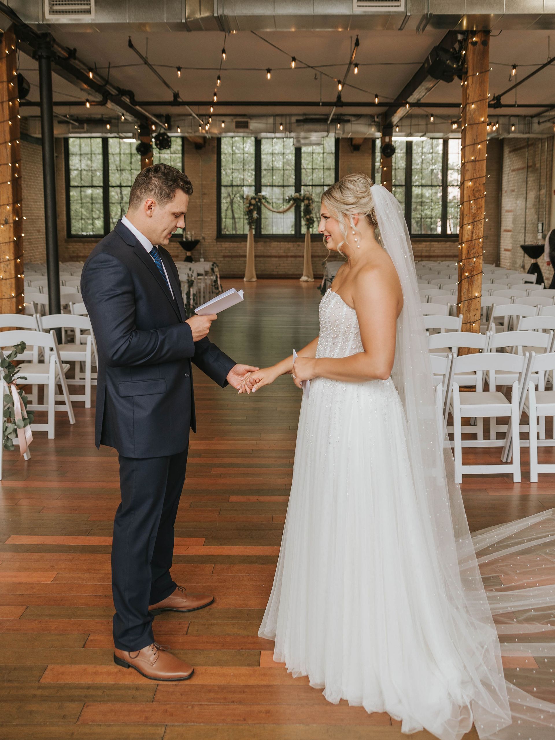 Wedding ceremony: Groom reading vows, holding bride's hand, indoor venue with wooden floor, white chairs, and string lights.