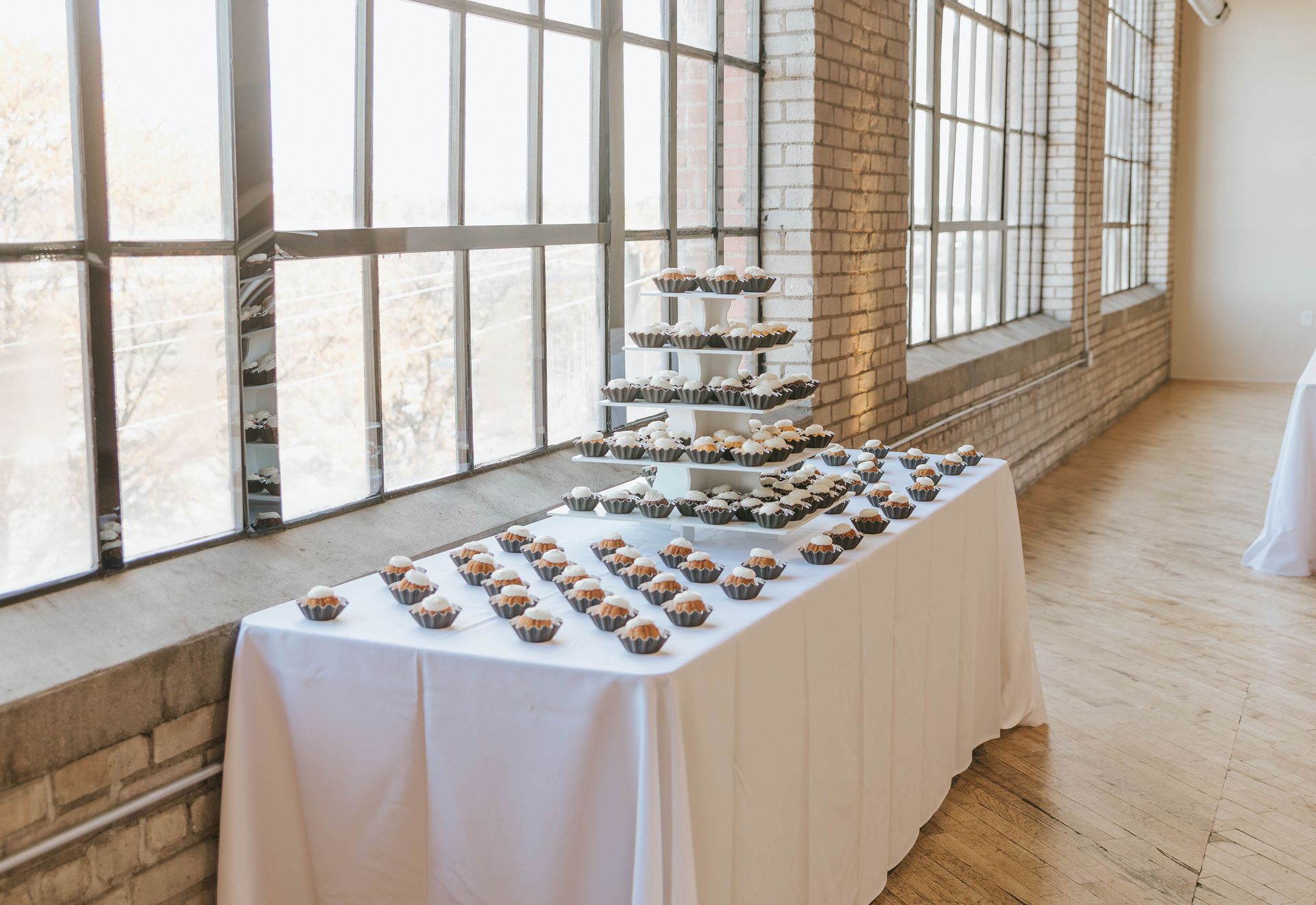 Cupcake display on a white-covered table near a large window. Tiered stand holds more cupcakes.