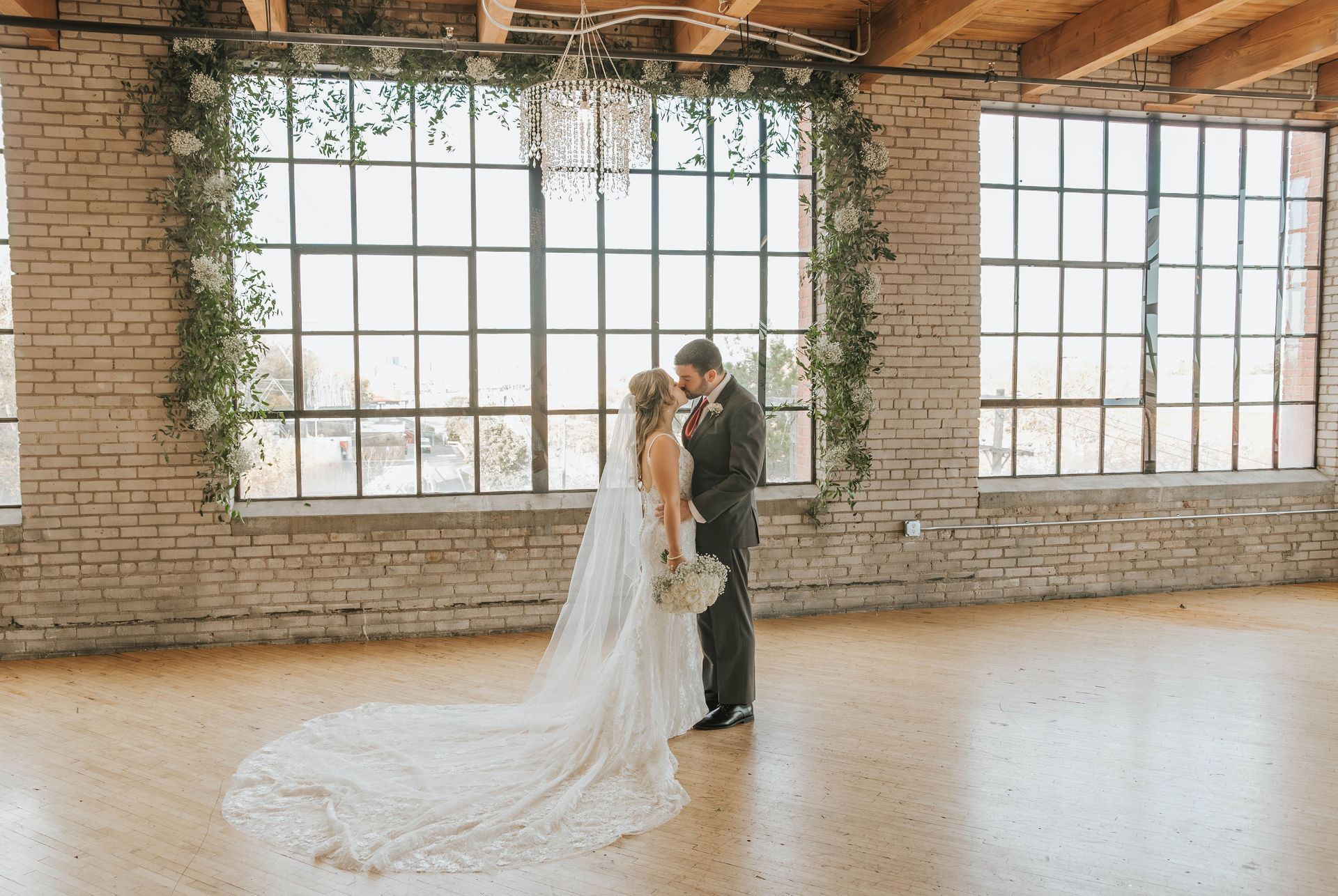 Couple kissing in front of large windows with floral decorations; bride in a wedding dress.