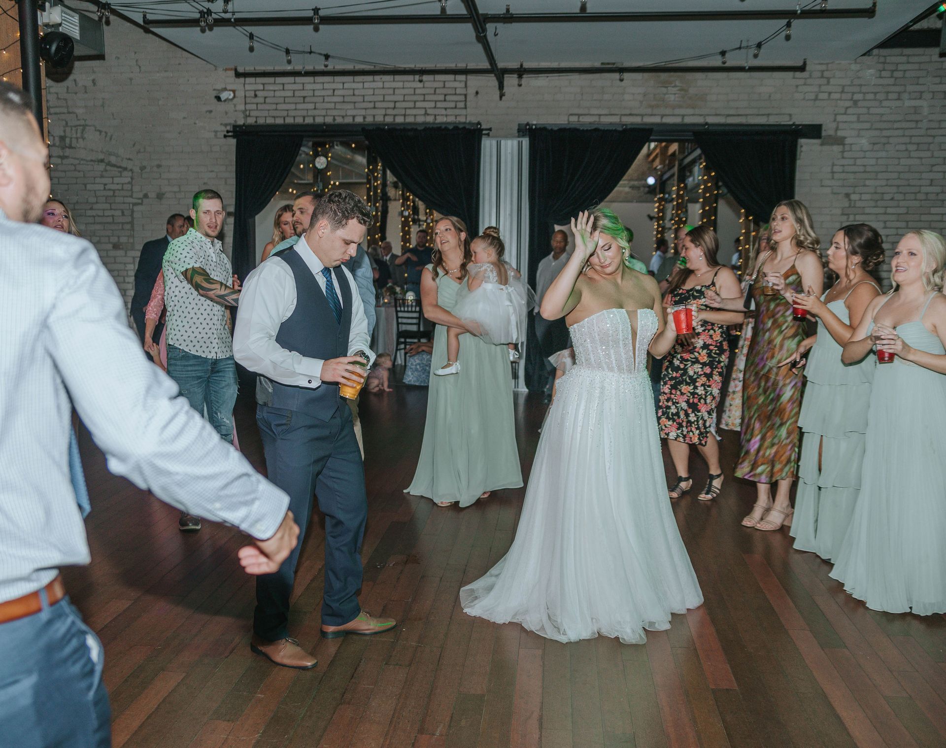 Wedding reception: Bride and groom dancing, surrounded by guests in a venue with dark curtains and wood floor.