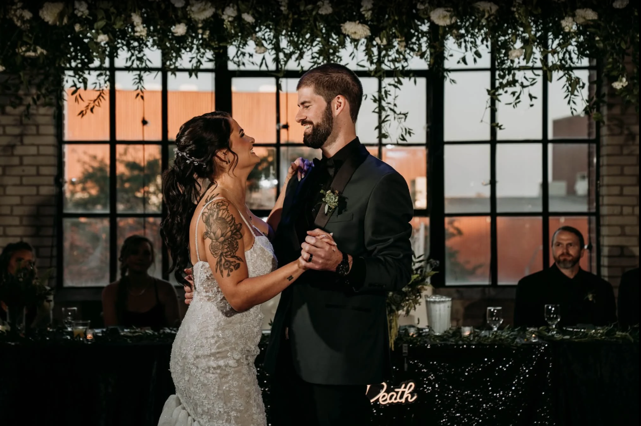 Bride and groom dance at reception; brick wall and large window in background.