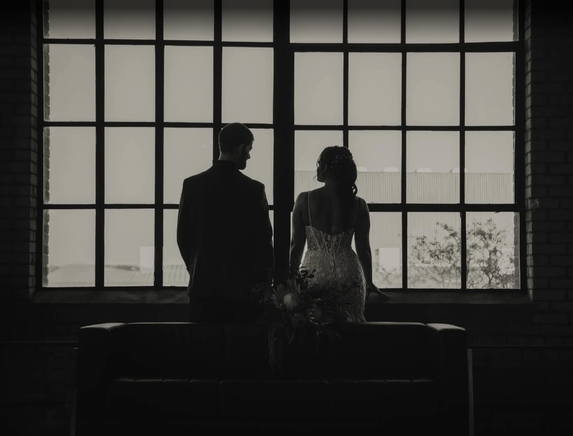 Couple stands by a large window, silhouetted. The bride wears a gown, the groom a suit.