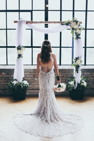 Bride in a beaded wedding dress, back to the camera, standing before an arch decorated with flowers and fabric.