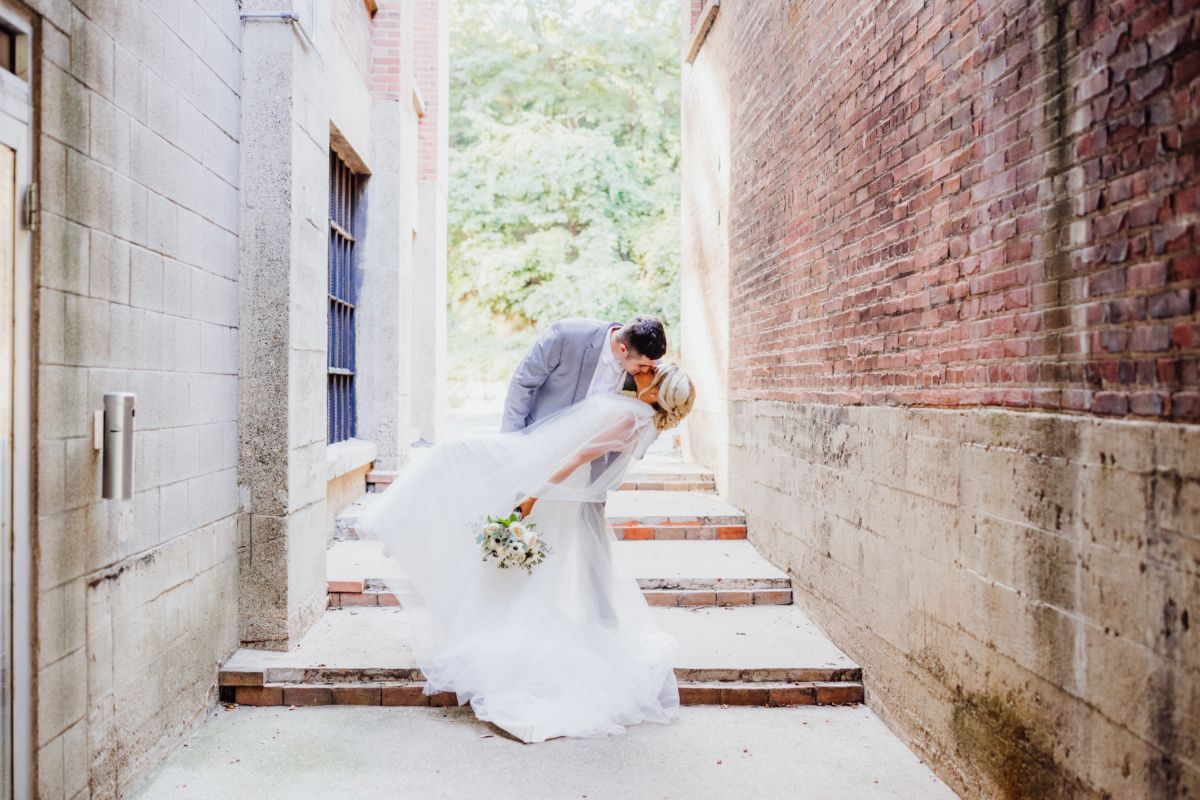 Groom dips bride for a kiss in a narrow alleyway with brick walls and stairs.