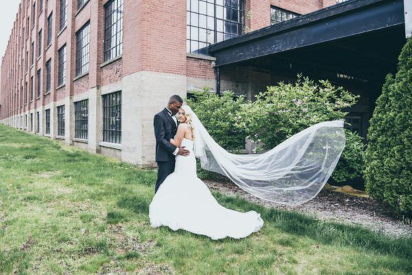 Wedding couple in front of brick building. Bride in white dress, veil blowing; groom in suit. Green grass.