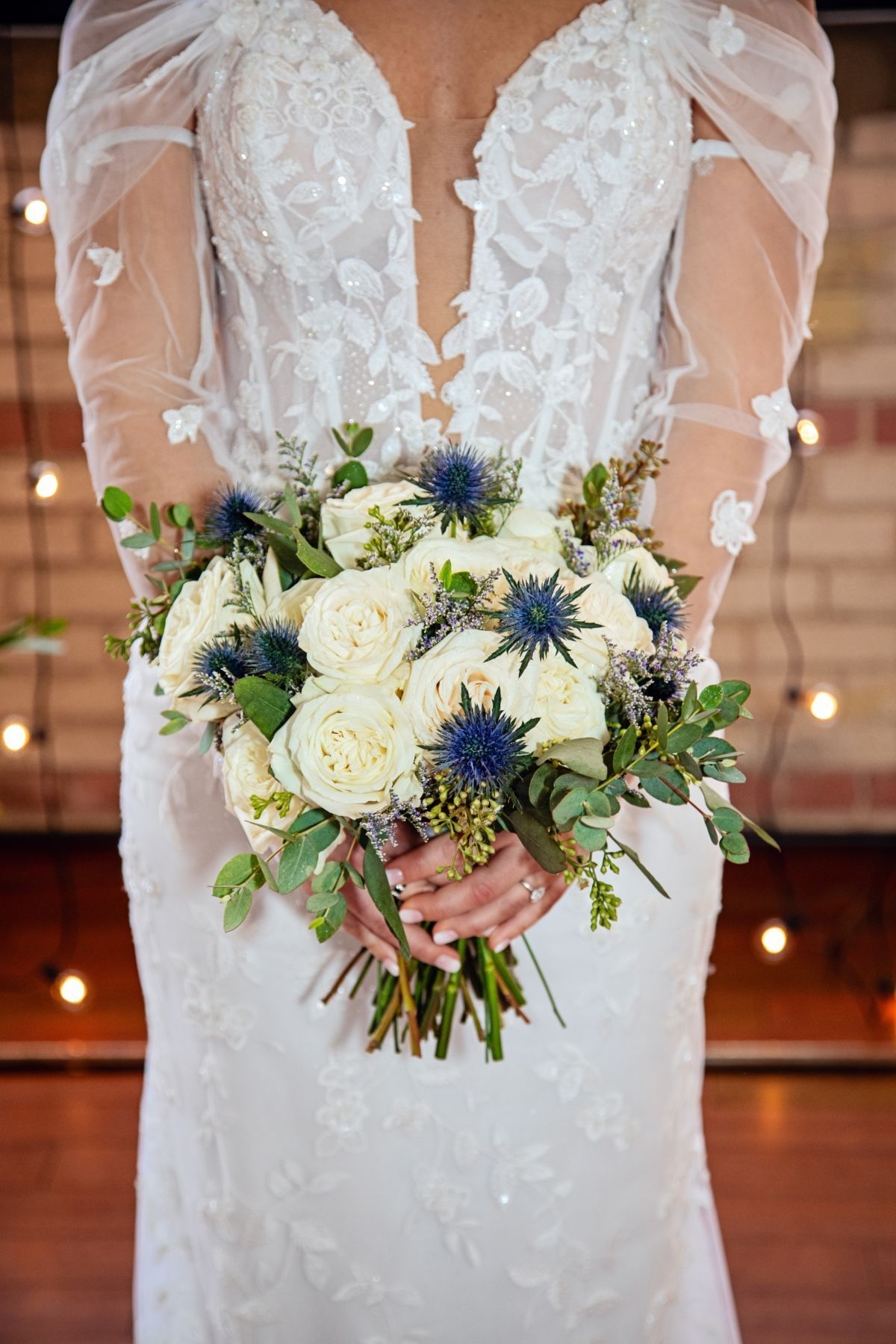 Bride in white lace dress, holding a bouquet of white roses and blue thistle.