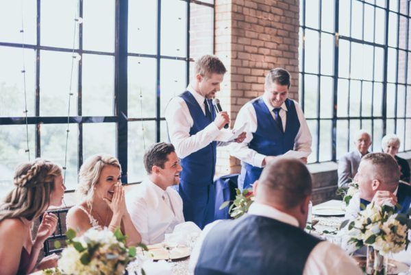 Two men in blue vests give a speech at a wedding. Guests seated at a table, some smiling.