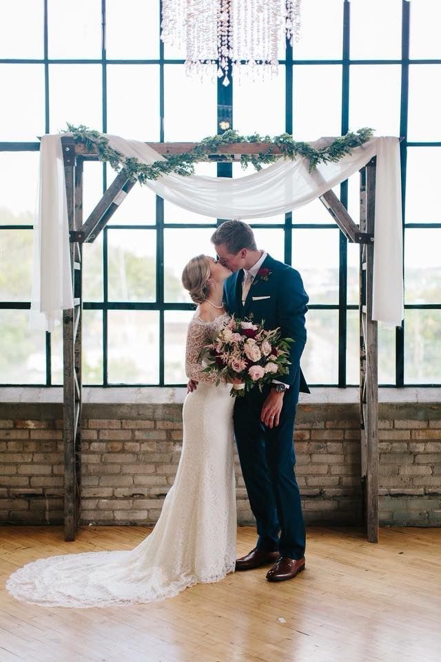 Couple kissing under a wedding arch, holding flowers. Arch is decorated with greenery and white fabric.