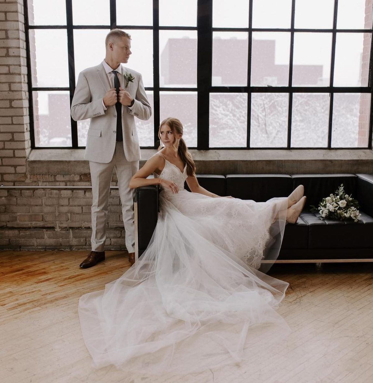 Bride in wedding dress reclines on a black couch, groom stands beside her. Window in background.