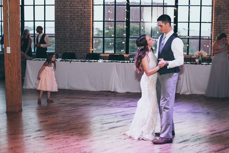 Newlyweds dance at reception, looking at each other. Flower girl watches nearby, other guests in background near windows.