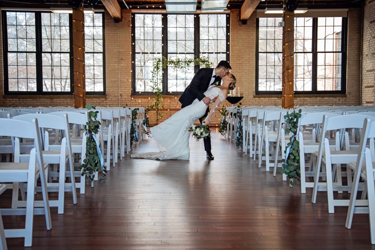 Groom dips bride in a wedding ceremony aisle. White chairs line the aisle, windows in the background.