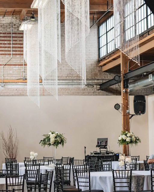 Wedding reception venue with white tablecloths, black chairs, and floral centerpieces. String lights hang from the ceiling.