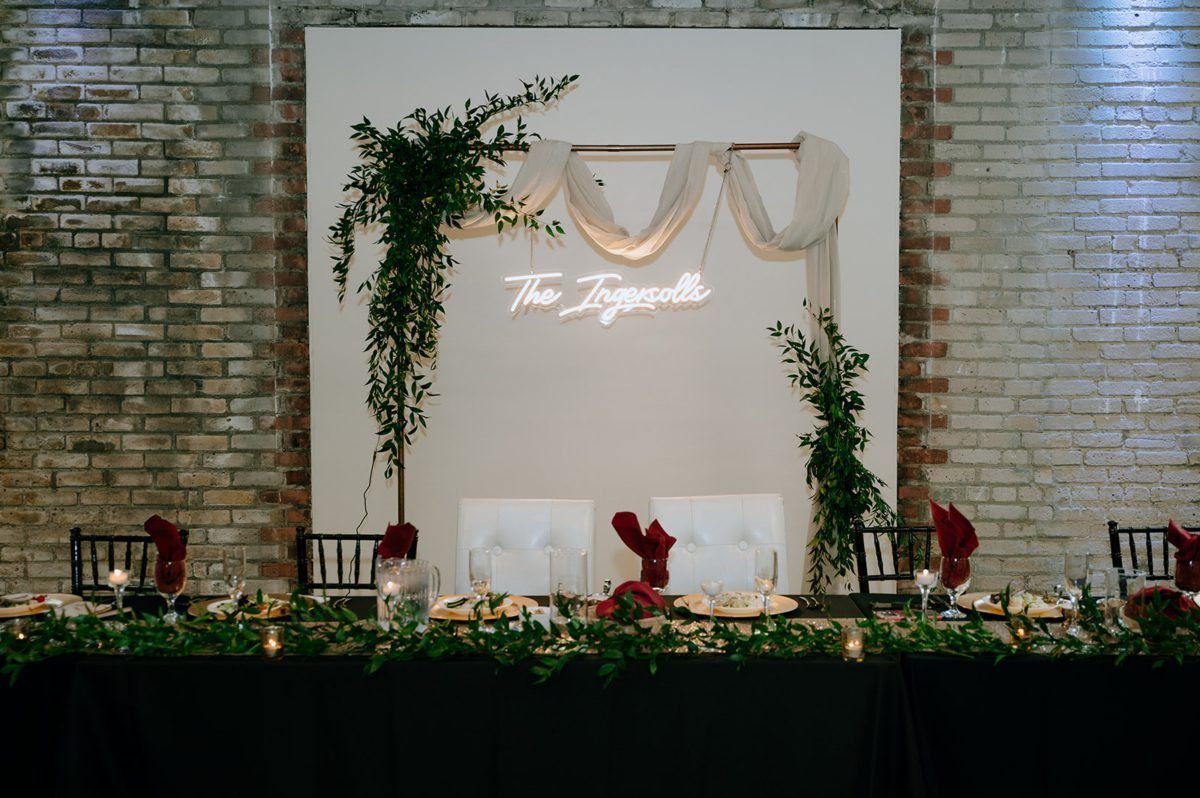 Wedding reception backdrop with neon sign, greenery, and draped fabric in front of a brick wall.
