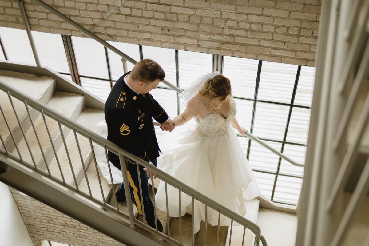 Couple in wedding attire descends a staircase. The groom wears a military uniform, bride a gown.